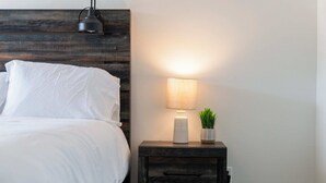 A neatly made bed with white linens, a bedside lamp, a small potted plant, and a dark wooden nightstand with drawers.