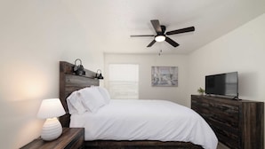 A bedroom with a bed, a ceiling fan, a TV, a lamp, and a painting on the wall. - Modern Poolside Apartment Near Gruene (New Braunfels)