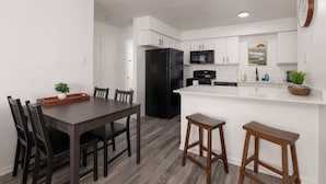 A modern kitchen with white cabinetry, a black refrigerator, a microwave, and a sink. There is a dining table with chairs and two bar stools. - Modern Poolside Apartment Near Gruene (New Braunfels)