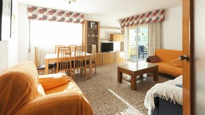 A living room with a sofa, a coffee table, a dining table with chairs, a television, and a kitchen area with cabinets and a refrigerator. - Acapulco Iv in Grau i Platja (Gandia)