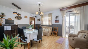 Un comedor con una mesa de vidrio, sillas y una araña. Hay un sofá y una butaca en la sala de estar, y un ventanal con cortinas.