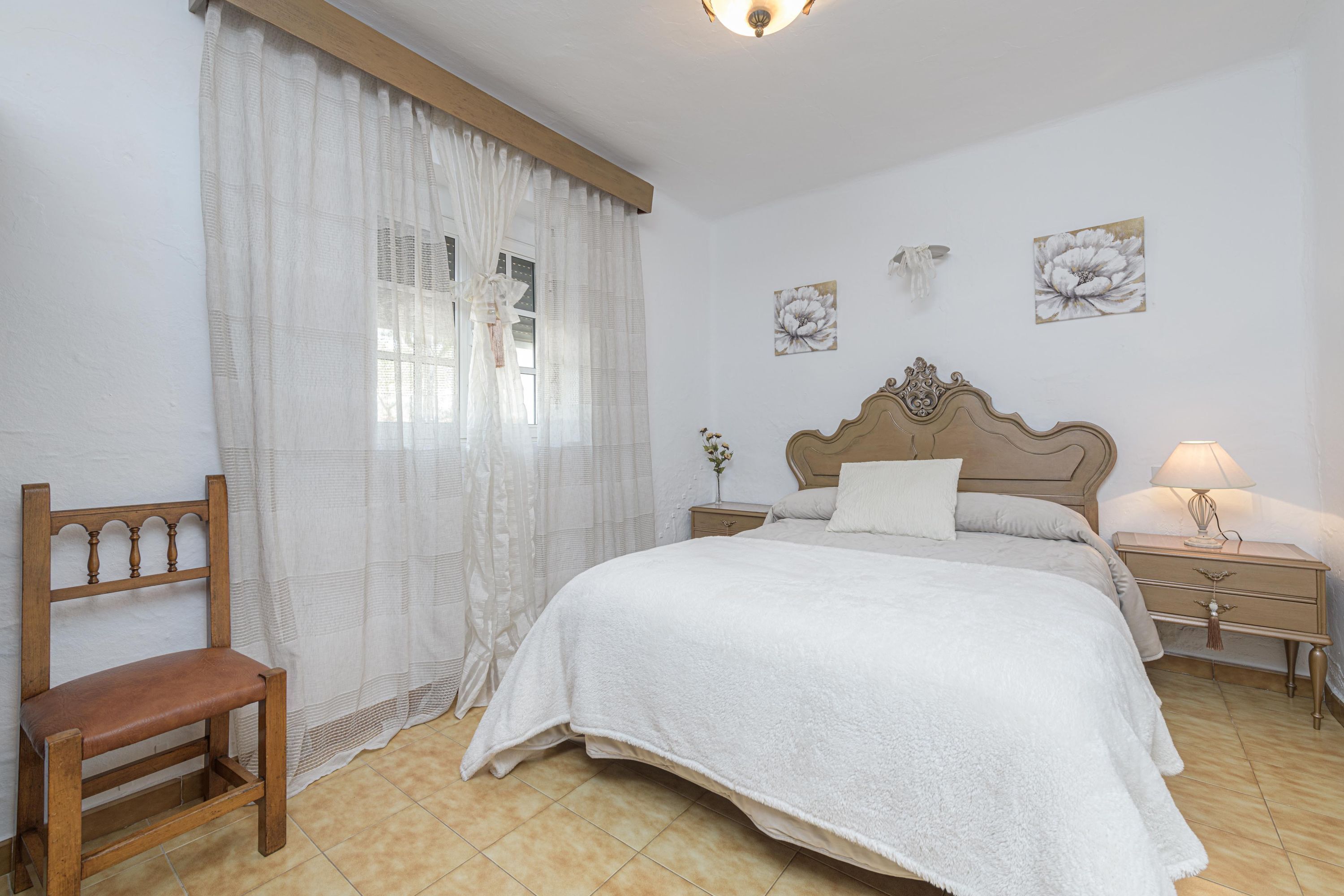 A bedroom with a bed, a wooden chair, a nightstand with a lamp, and a window with sheer curtains.