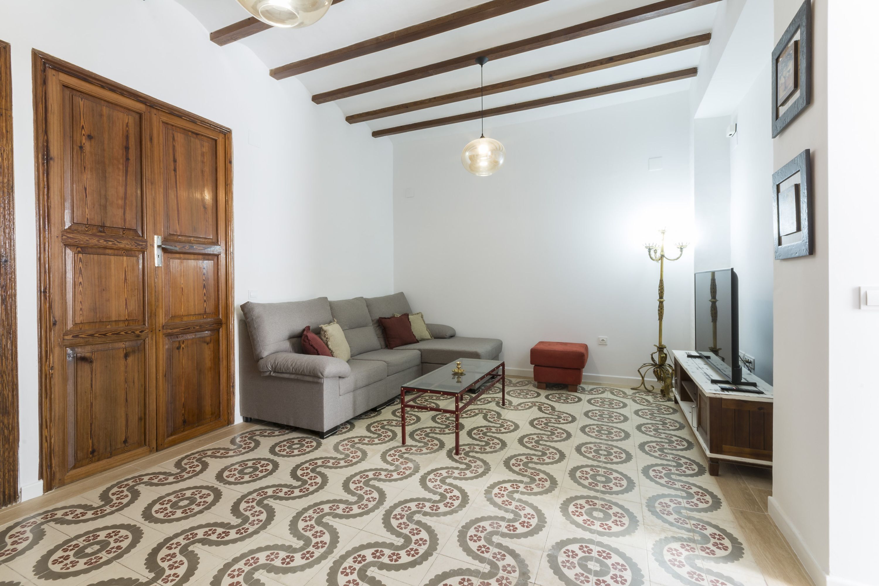 Un soggiorno con divano, tavolino e pavimento decorato. Si vede un armadio in legno, un mobile TV e un lampadario.