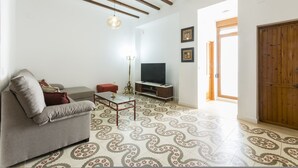 Un salon comprenant un canapé, une table basse et un téléviseur, des poutres en bois au plafond, des carreaux de sol à motifs et une porte en bois.
