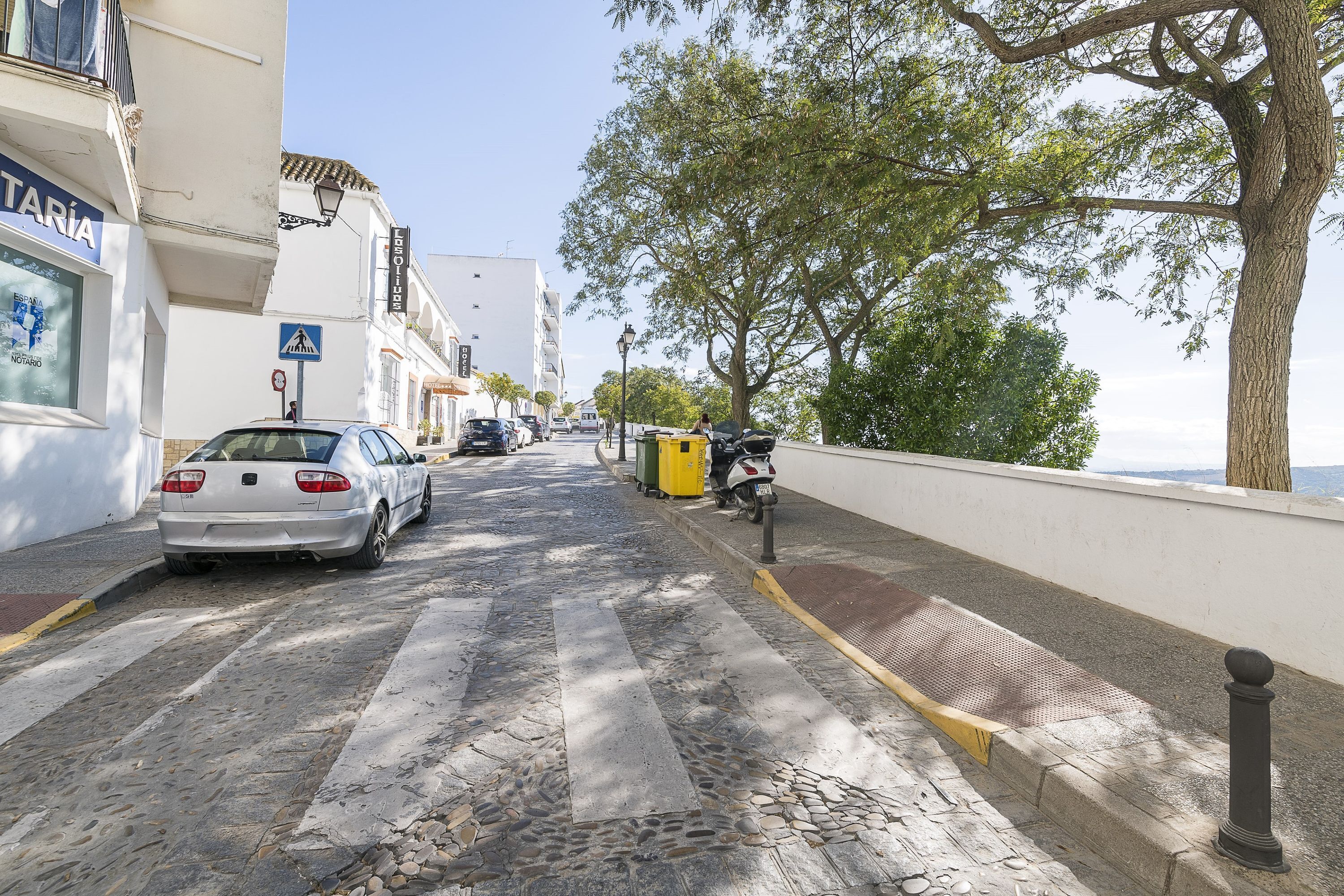 Een straat met kinderaardewerkstenen, met een witte auto aan de linkerkant, een gele prullenbak en een scooter aan de rechterkant.