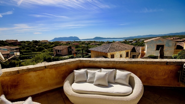 Ein Balkon mit Rattan-Sofa und Kissen mit Blick auf ein Küstendorf mit traditionellen Häusern und einem strahlend blauen Himmel.