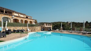 Ein Schwimmbecken mit Liegestühlen und Sonnenschirmen, ein Gebäude aus Stein mit Bögen und Blick auf einen Hügel.