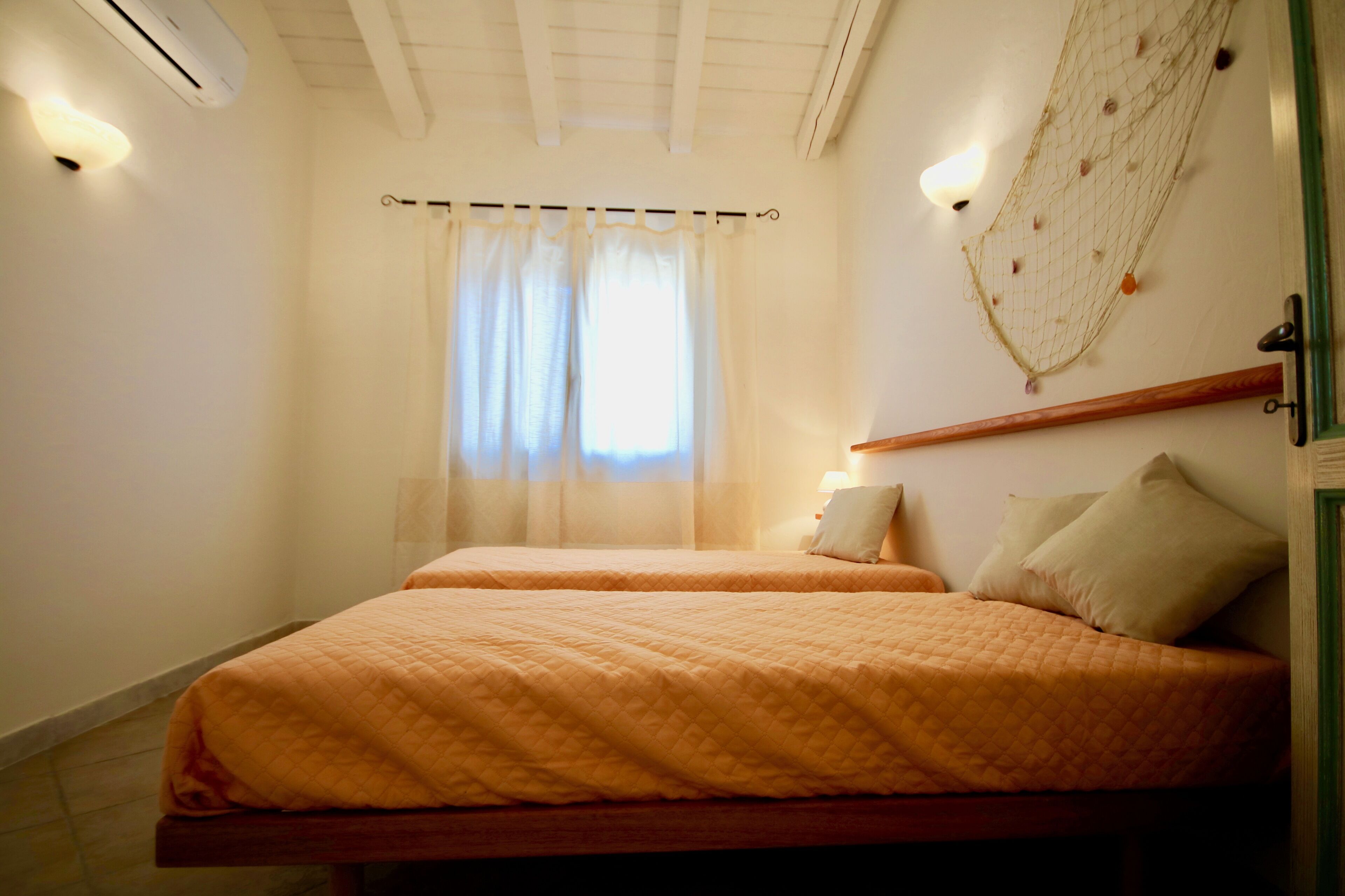 A bedroom with two beds, a window with sheer curtains, a wall-mounted light fixture, and a decorative net on the wall.