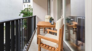 A balcony with a wooden table and two chairs, a potted plant, and a window with a view of buildings and greenery.