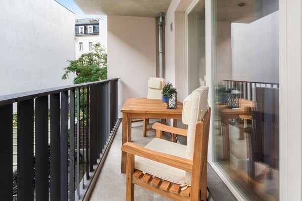 A balcony with a wooden table and two chairs, a potted plant, and a window with a view of buildings and greenery.