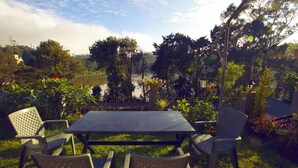 Eine Terrasse im Freien mit Tisch und Stühlen, umgeben von Grünpflanzen und Blick auf einen See.