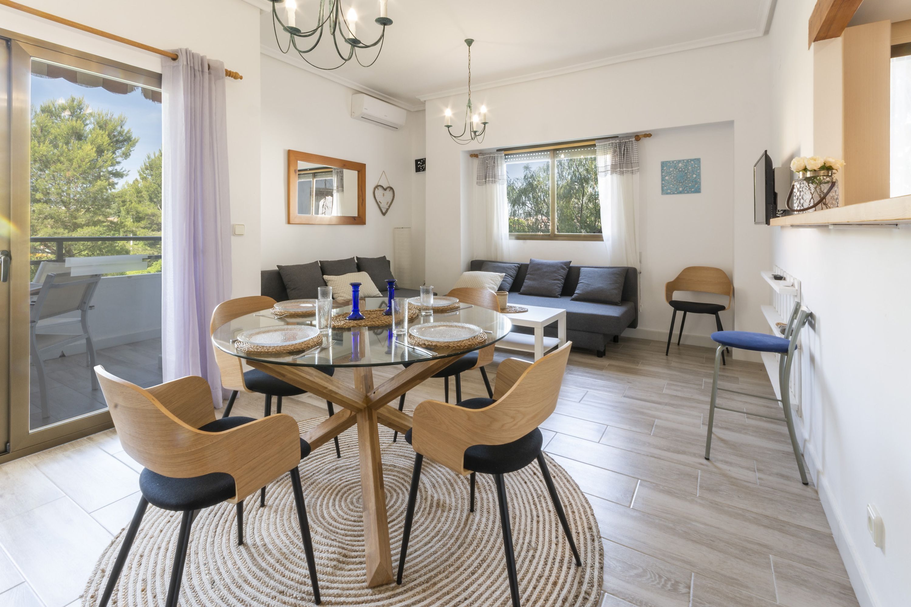 A dining area with a glass table, wooden chairs, and a round rug. There is a sofa and a chair in the background.