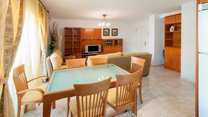 A living room with a glass-top dining table, wooden chairs, a sofa, a TV, wooden shelving, and a door leading to another room. - Cascades in D nia With 2 Bedrooms and 1 Bathrooms (Denia)