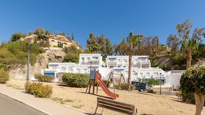 Un parque infantil con tobogán y columpios, un banco y una calle frente a un edificio blanco con puertas azules.