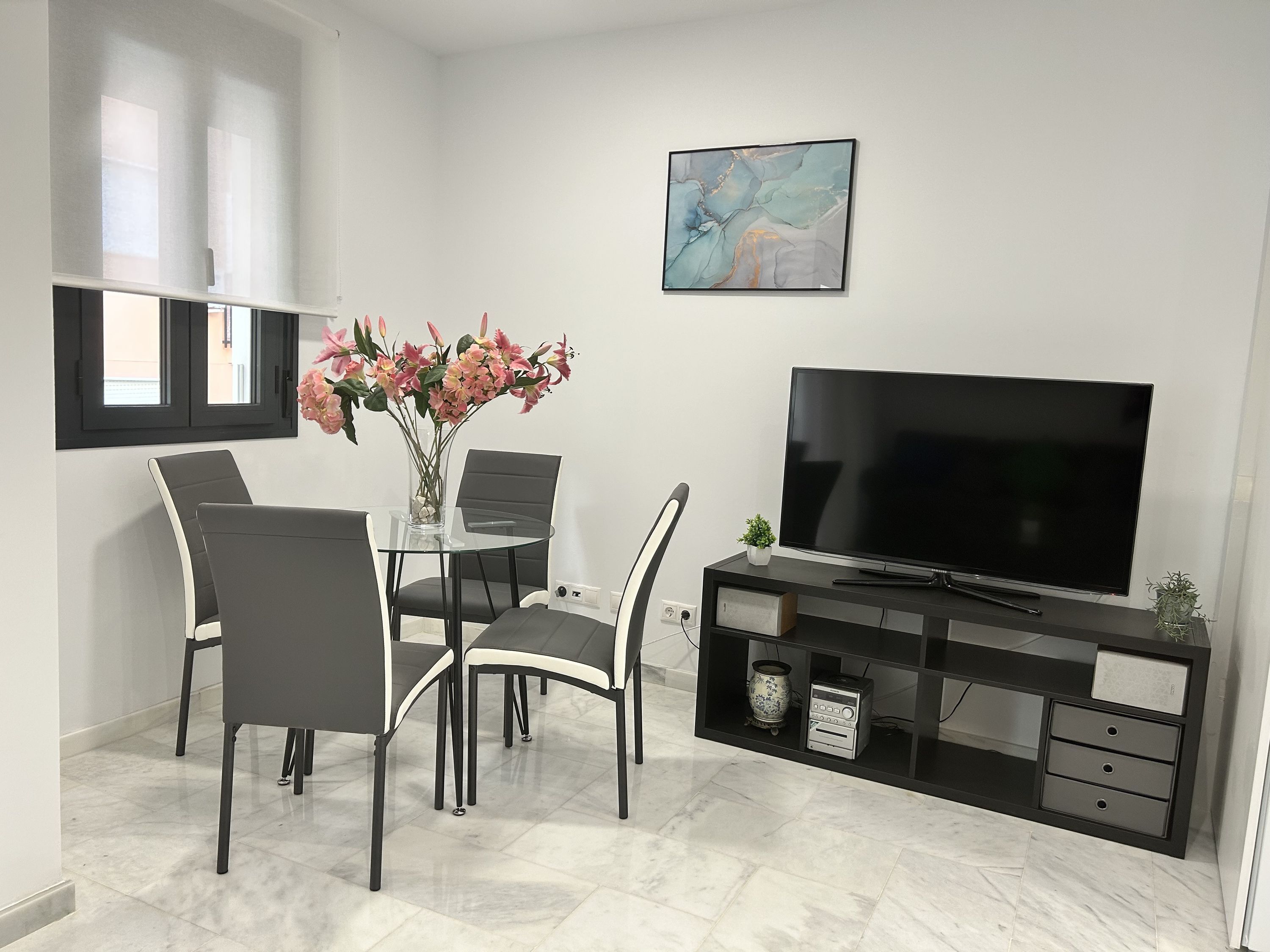 Un soggiorno moderno con un tavolo da pranzo in vetro, quattro sedie, un televisore piatto, un mobile TV nero e un vaso con fiori rosa.