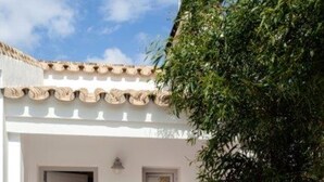 A white building with a tiled roof, a door, and a bench with cushions. There are potted plants and a table with chairs outside. - Casa Tecolote in Chiclana de la Frontera (Chiclana de la Frontera)