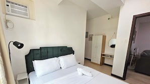Een net opgemaakt bed met witte lakens, een donkergroen bekleden hoofdbord en een witte nachtlamp.