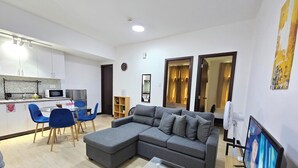Een moderne woonkamer met een grijze modulaire bank, een glazen salontafel en een televisie. Er is een eethoek met een witte tafel en blauwe stoelen. De keuken is zichtbaar, met een magnetron en een koelkast.