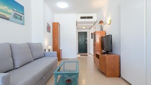 Una sala de estar con sofá, una mesa de centro de vidrio, un televisor sobre un mueble de madera y una puerta al fondo.