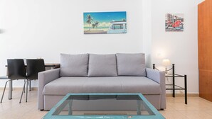 Una sala de estar con un sofá gris, una mesita de centro color turquesa y dos sillas negras. Hay dos cuadros colgados en la pared, uno con una escena de playa y otro de una bicicleta.