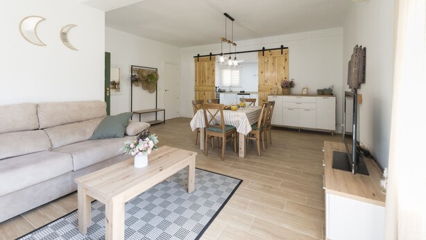 Een moderne woonkamer met een bank, salontafel en een eethoek met stoelen en een tafel. Achtergrond toont een keuken met houten kasten en een hangende lamp.