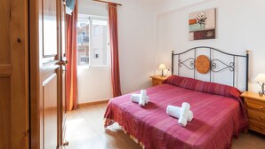 Un letto rifatto con una coperta rossa e due asciugamani arrotolati, un comodino con una lampada, e una finestra con tende.