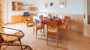 Un accogliente soggiorno con un tavolo da pranzo per quattro persone, circondato da sedie in legno. Una televisione è posata su un mobiletto in legno, e sopra si trovano delle mensole con oggetti decorativi.