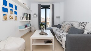Un salon moderne avec un canapé blanc, une table basse blanche et un téléviseur fixé au mur. Des objets décoratifs sont disposés sur des étagères, et un vase rempli de fleurs se trouve sur la table.