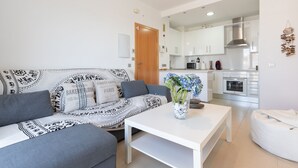 Un salon moderne avec un canapé gris, une table basse blanche et un vase de fleurs. On aperçoit une cuisine à l’arrière-plan, avec des meubles blancs et des appareils électroménagers en acier inoxydable.