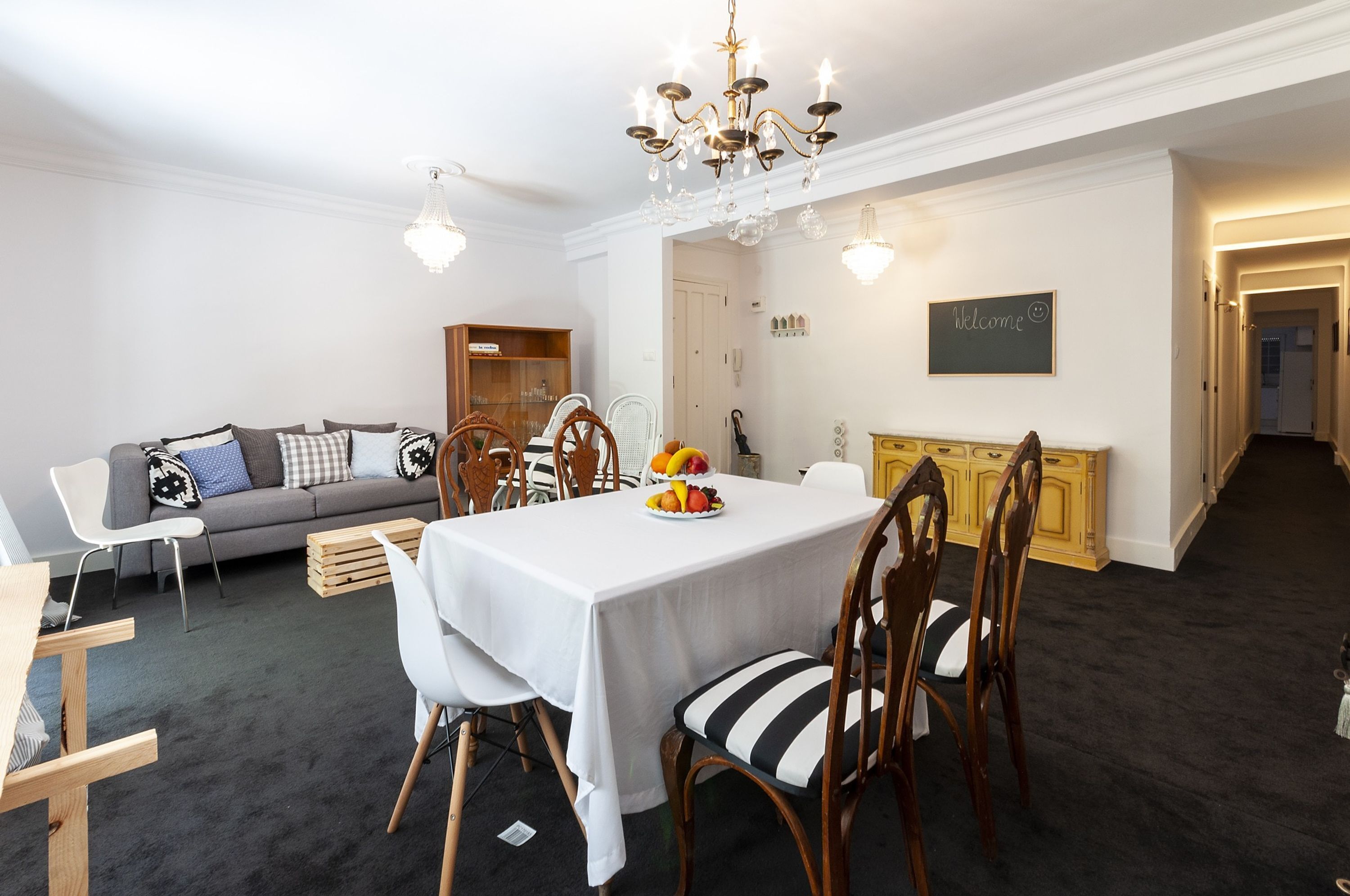 A dining area with a white table, striped chairs, and a fruit centerpiece. A grey sofa and wooden cabinet are in the background.
