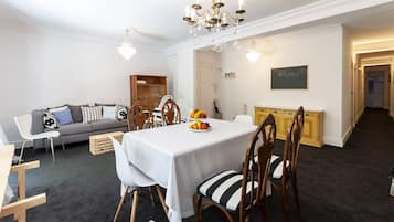 A dining area with a white table, striped chairs, and a fruit centerpiece. A grey sofa and wooden cabinet are in the background.