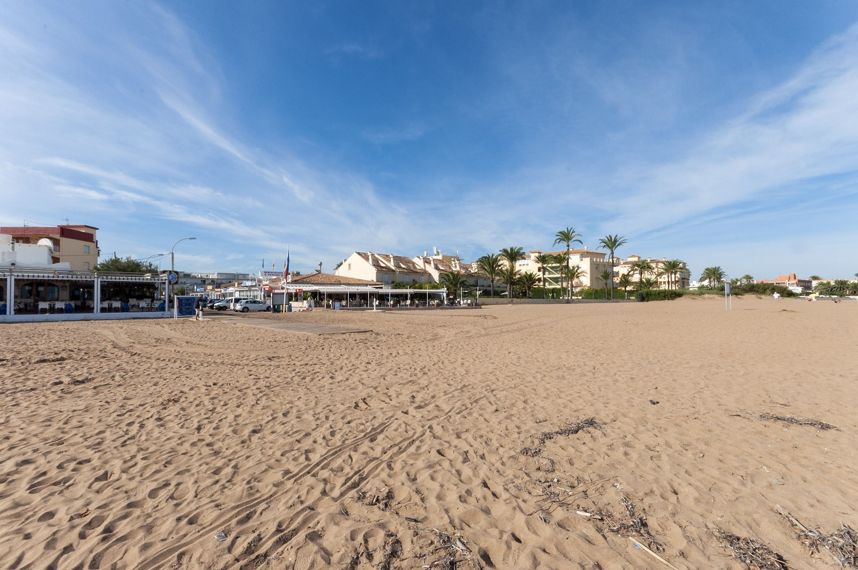 A wide sandy beach with buildings in the background, including a structure with a red roof and a covered area.