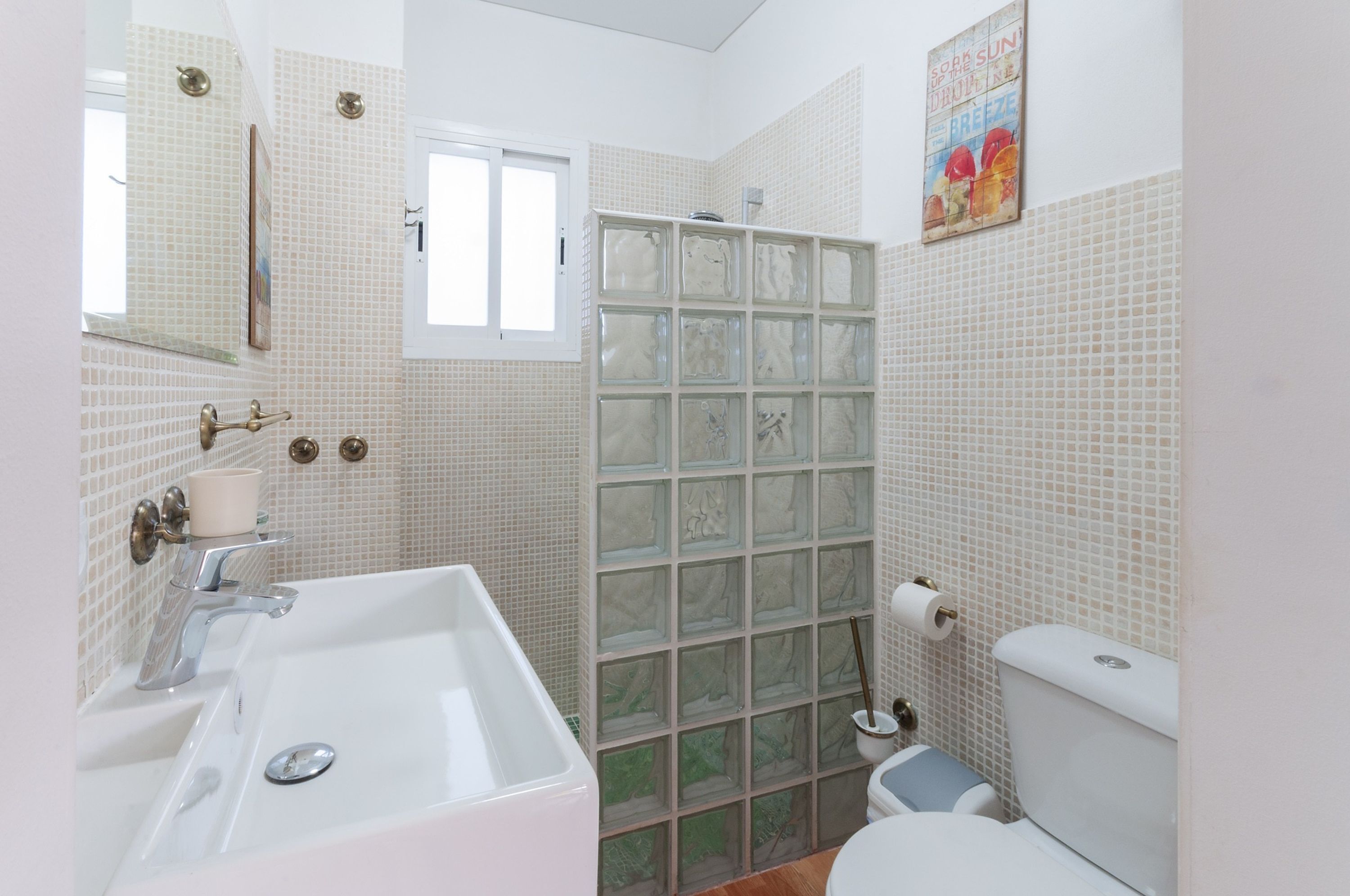 Une salle de bain avec un lavabo blanc, un miroir et des toilettes. Il y a une fenêtre et une douche avec une porte en verre.