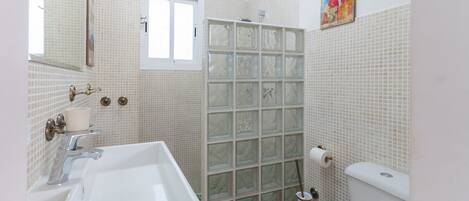 Une salle de bain avec un lavabo blanc, un miroir et des toilettes. Il y a une fenêtre et une douche avec une porte en verre.