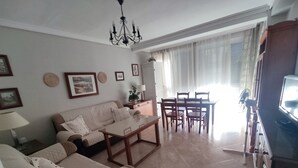 A living room with a sofa, a coffee table, a dining table with chairs, a chandelier, and a television.