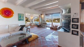 Zona d'estar àmplia amb un sofà blanc, una taula de centre de vidre i una obra d'art colorida de grans dimensions a la paret. Hi ha una televisió sobre un suport i una llar de foc. La habitació té un paviment de rajoles i vista a l'exterior a través de grans finestres.