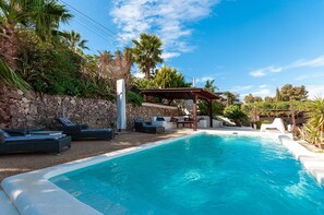 Une piscine dotée de chaises longues et d’une pergola, entourée de verdure et d’un mur de pierre.