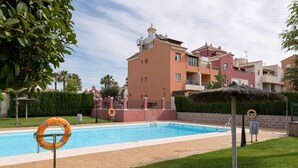 Una piscina con un flotador salvavidas, una sombrilla de paja y un edificio con balcones al fondo.