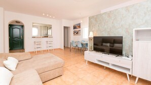 Una sala moderna con un sofá beige, un televisor sobre un mueble blanco y un comedor con sillas y una mesa.