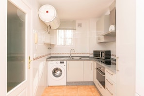 Une cuisine compacte équipée d’un lave-linge, d’un four à micro-ondes, d’un four traditionnel et d’une fenêtre avec des stores.