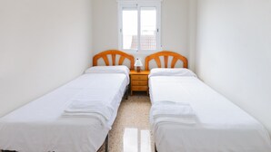 A small, clean room with two single beds, a wooden nightstand, a white lamp, and a window with a view of buildings. - Turia in Tavernes de la Valldigna (Tavernes de la Valldigna)