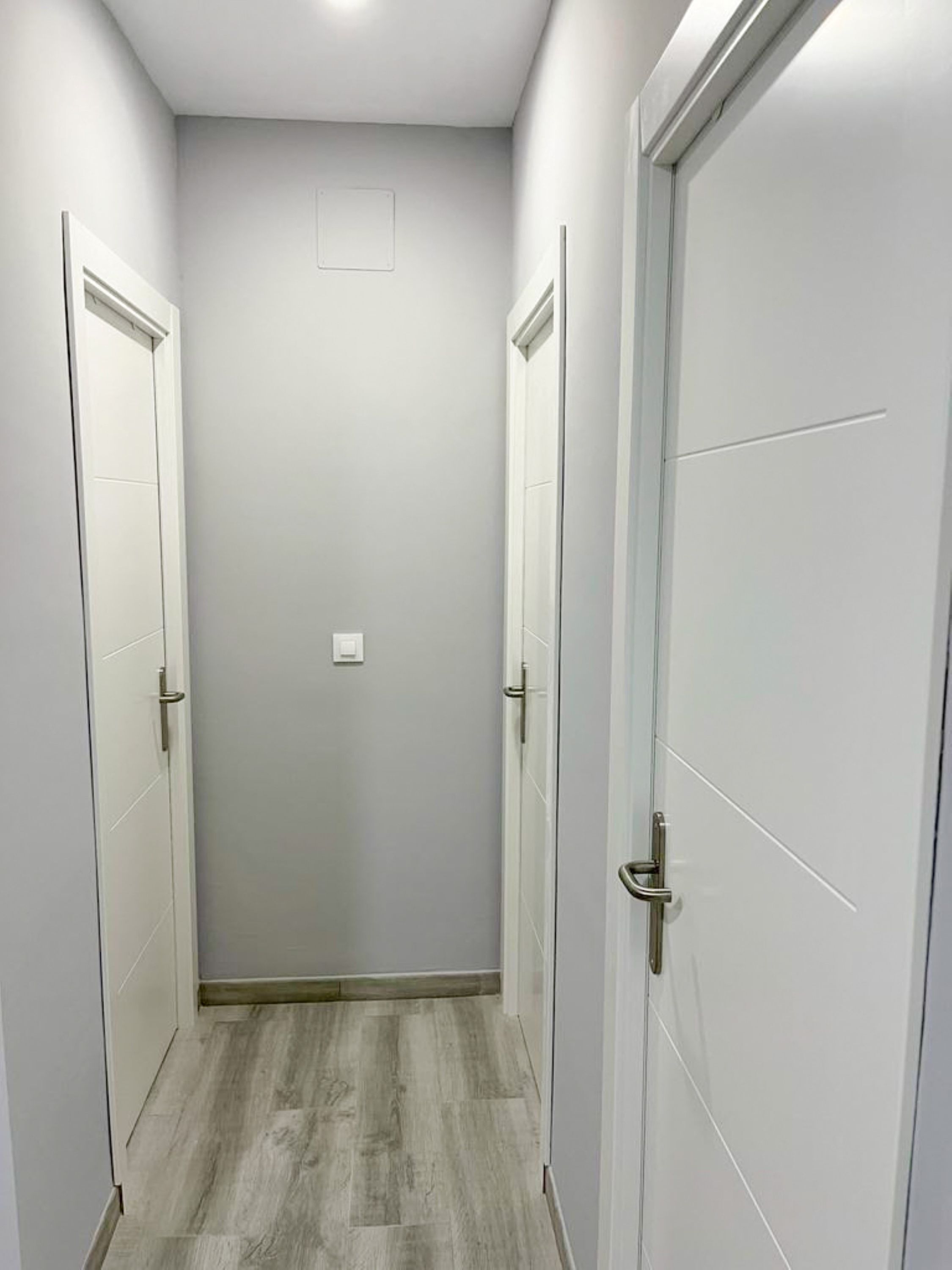 A narrow hallway with two doors on either side, a light switch on the wall, and a wooden floor.
