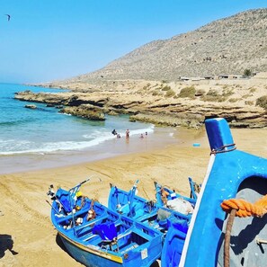 A beach with rocky terrain and a few people in the water. There are several blue boats lined up on the sand.