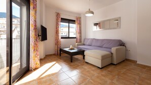 A living room with a sofa, a coffee table, and a TV. There is a window with curtains, a ceiling light, and a mirror on the wall. - Tolox 2 in Tolox With 1 Bedrooms and 1 Bathrooms (Tolox)