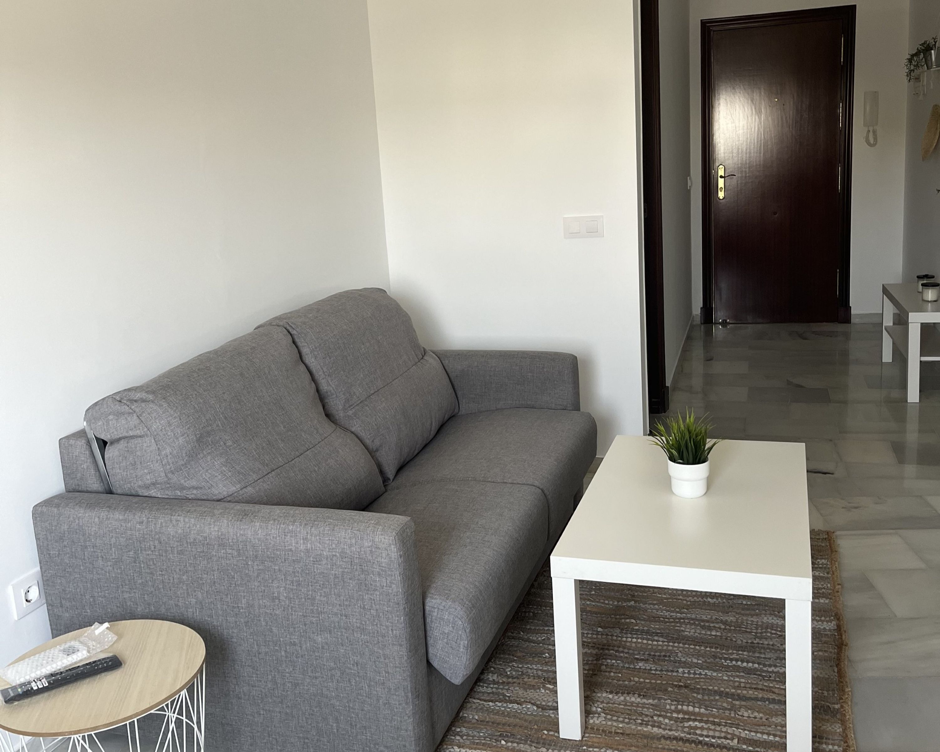 Un salón con un sofá gris, una mesa de centro blanca y una mesita redonda con un control remoto.