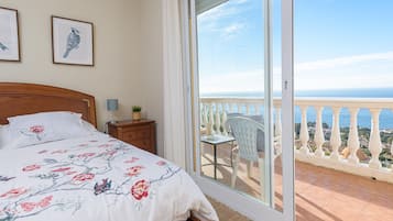 A bedroom with a bed, a nightstand, a chair, and a balcony with a view of the sea.