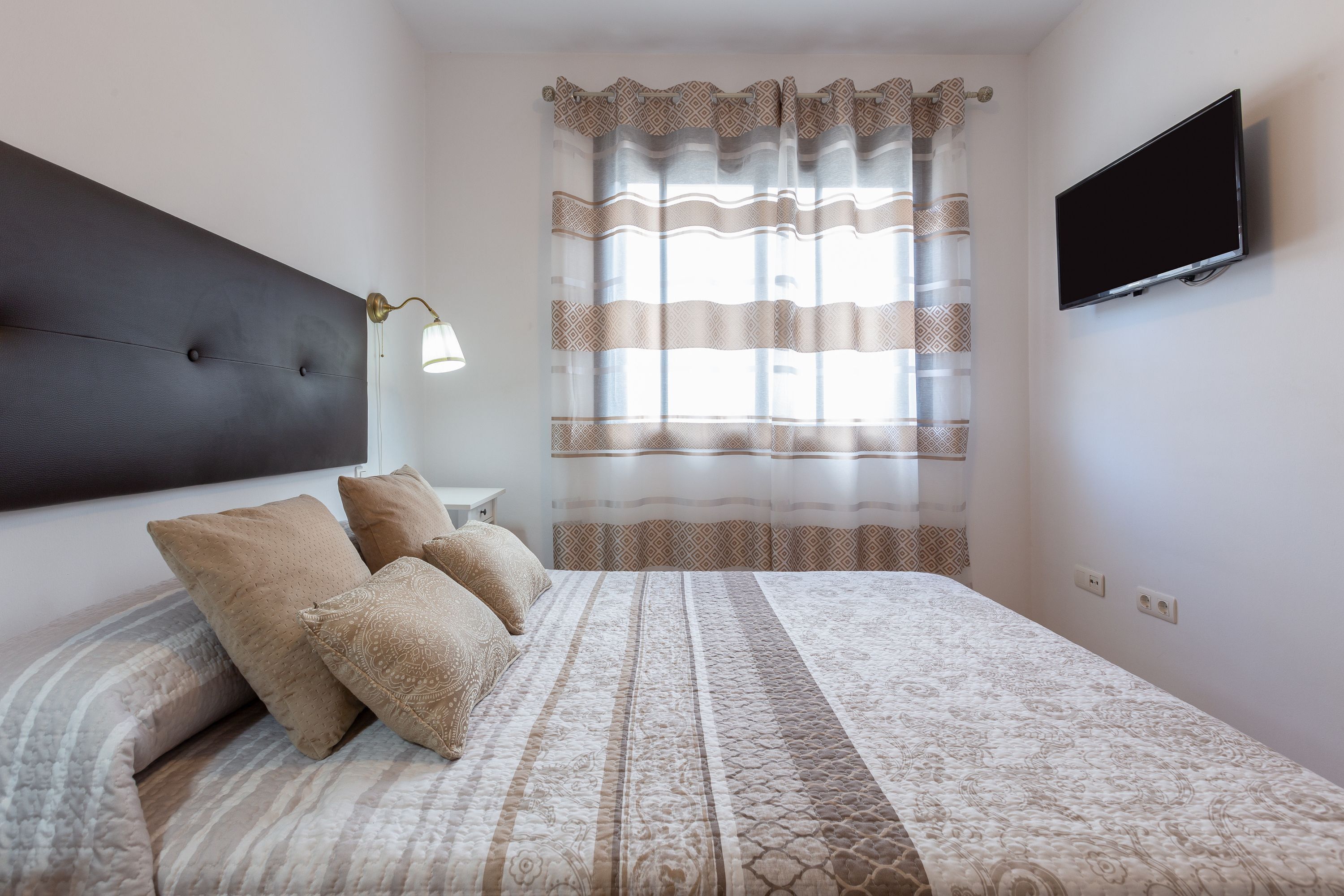 Een slaapkamer met een bed, kussens, een bedlampje, een televisie en een raam met gordijnen.