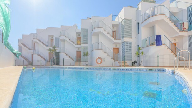 A swimming pool with a clear blue water surface, surrounded by white buildings with balconies and a staircase leading to the upper floors. - Villablanca Home in Villablanca (Villablanca)