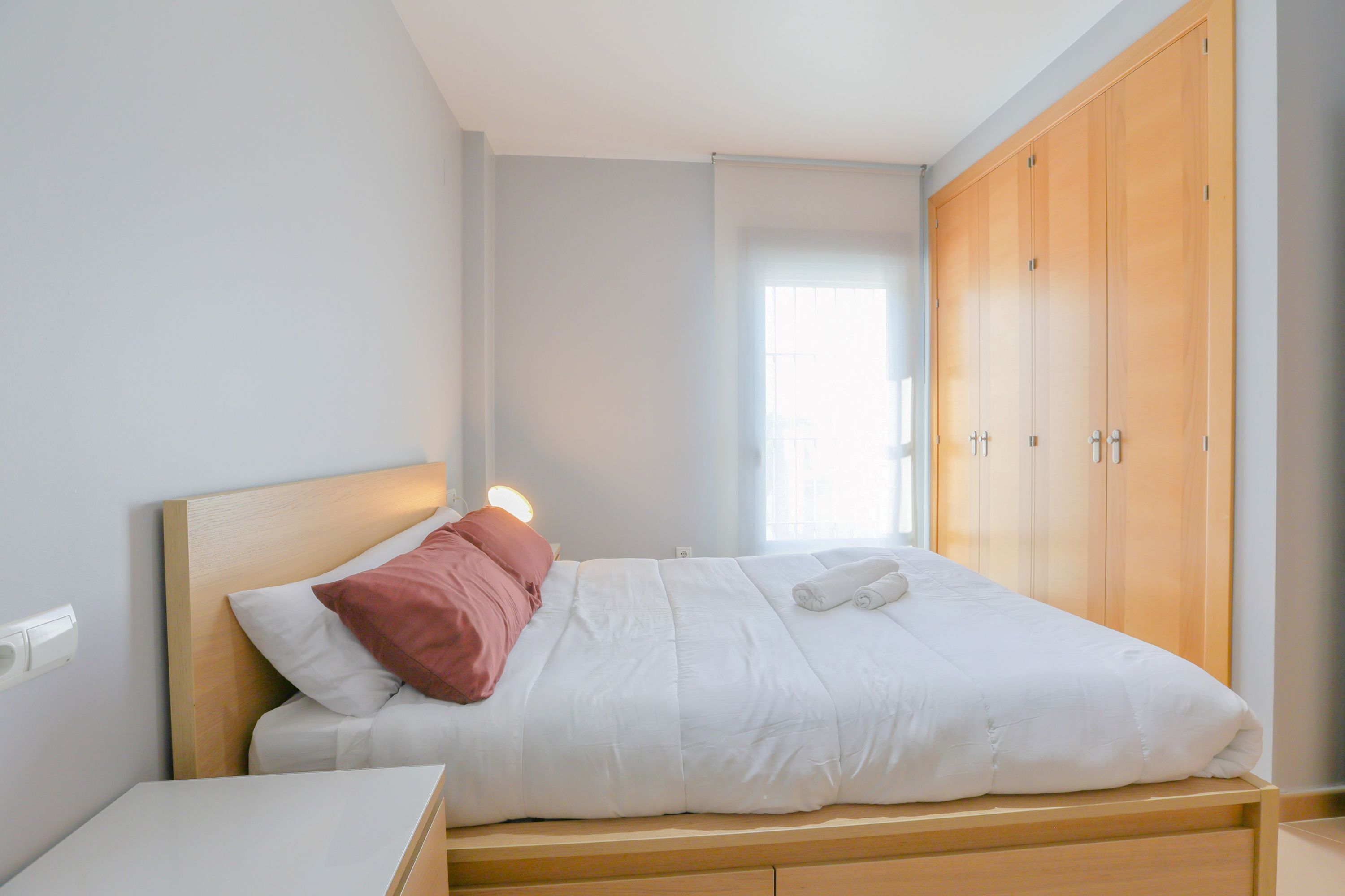 Ein ordentlich bezogenes Bett mit weißer und pinkfarbener Bettwäsche, ein hölzerner Kopfteil und ein Holzschrank in einem hellen Raum mit Fenster.