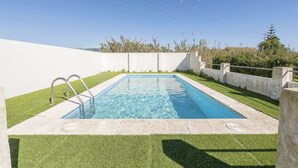 Una piscina rectangular con escaleras de metal, rodeada por un suelo de hormigón y césped artificial verde.